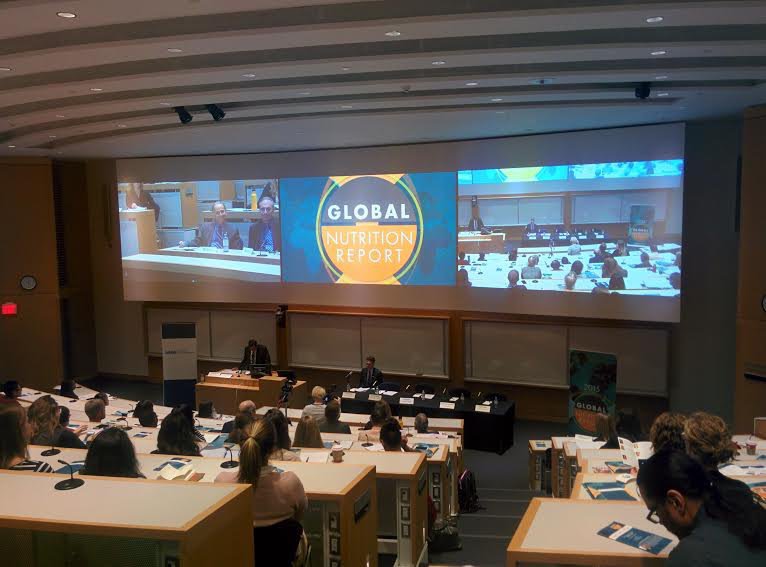 Delegates sat desks on a tiered platform, viewing a panel and speaker in front of large wide screen presentation projection at Global Nutrition Report 2015 Event Toronto